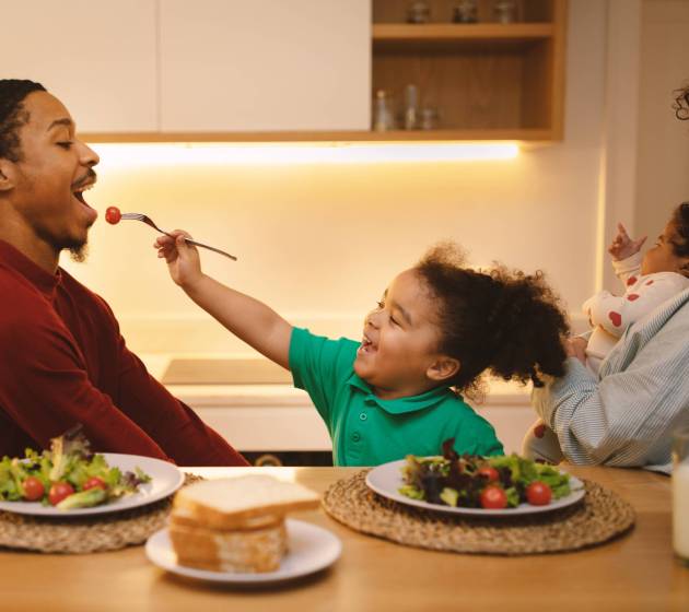 Beautiful happy loving interracial family have lunch at modern kitchen at home. Cute baby girl feeding her dad african american man, hispanic curly woman holding infant, enjoying time together