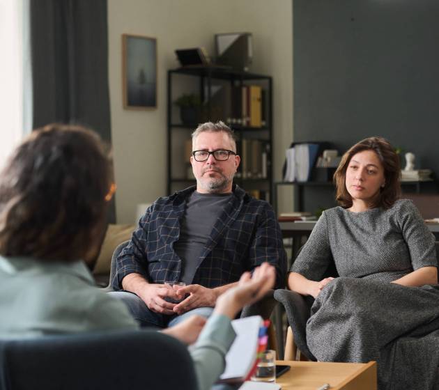 Couple sitting in office discussing relationship issues with therapist holding clipboard. Room has bookshelf, lamp, abstract painting, and large window providing natural light