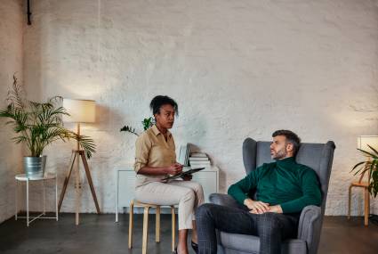 African female psychiatrist writing information of middle age male patient while sitting in armchair and talking about his illness