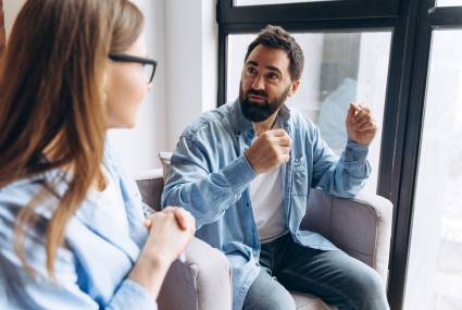 Bearded man talking to psychologist sitting on armchair in front of window during therapy session