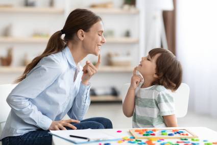 Speech training for kids. Professional woman specialist training with little boy at cabinet, teaching him right articulation exercises, side view
