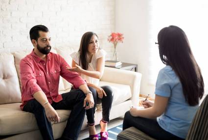 Angry woman pointing at her husband and discussing with psychologist. Unhappy couple talking to marriage counselor.